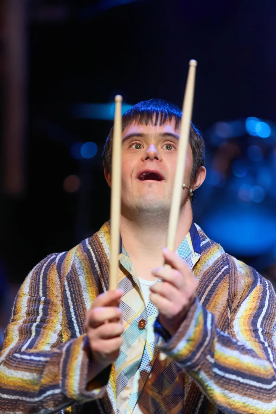 An actor in a striped dressing gown holds up a pair of drumsticks under blue stage light
