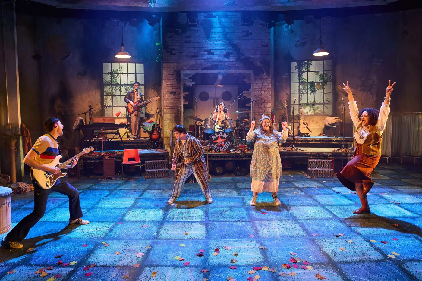 The full company on a tiled stage: guitarists, a drummer and three performers dancing with their arms raised